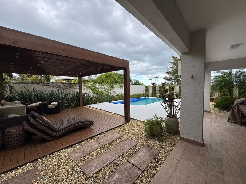 Deck de madeira com sofás sob o pergolado e a piscina ao fundo