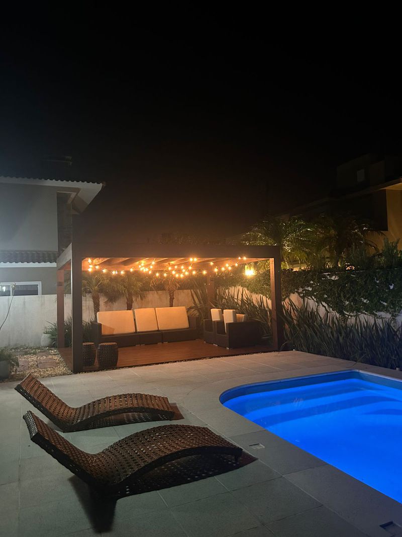 Pergolado iluminado com luzes de varal, espreguiçadeiras e piscina acesa à noite