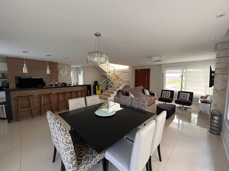 Sala de estar e jantar integradas à cozinha, com lareira, piso claro e escada ao fundo